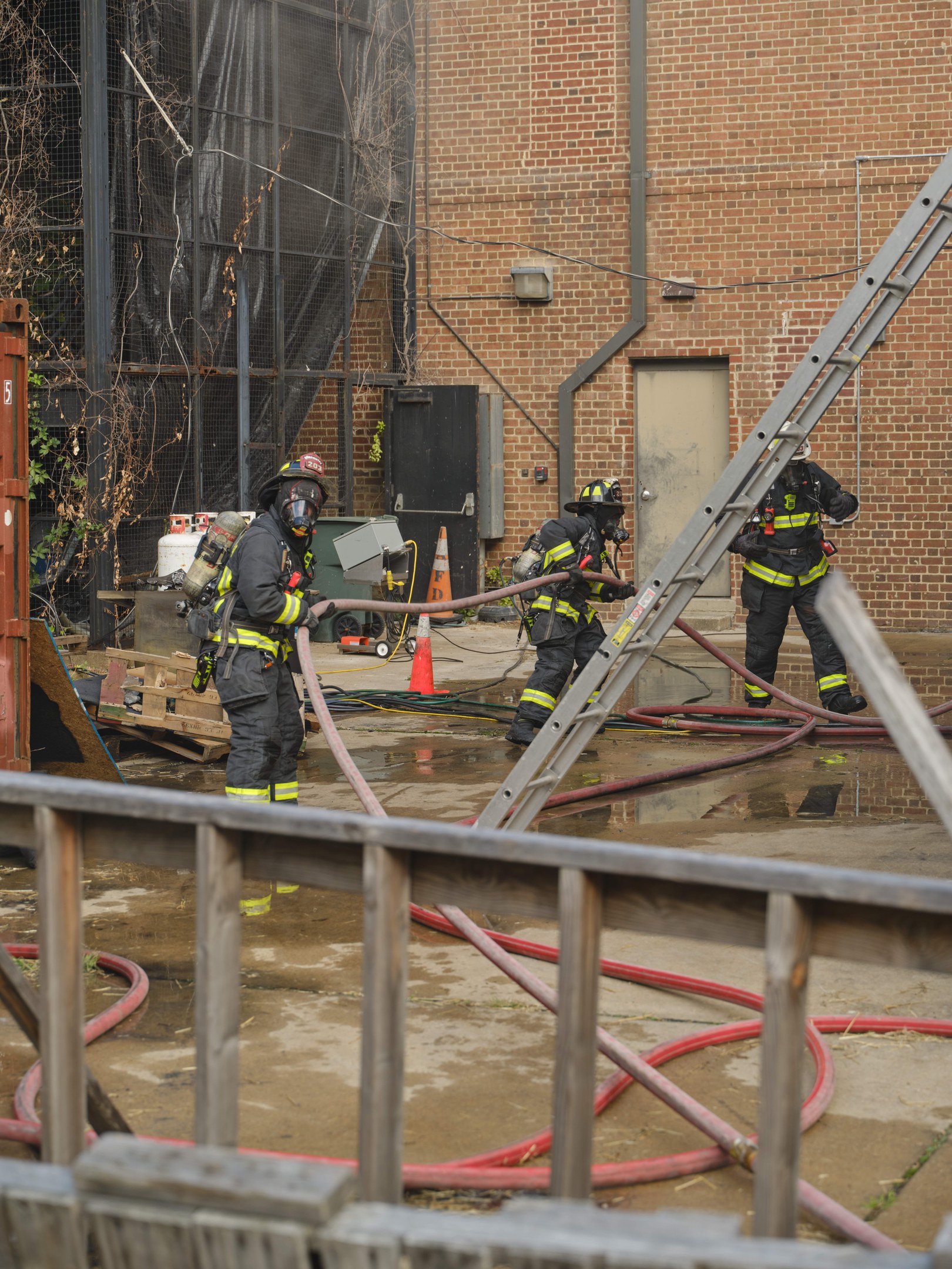 Feuerwehrleute in Helmen arbeiten daran, ein Gebäude Feuer zu löschen, während sie Schläuche halten, mit einem Metallzaun, Rohren, einem Container, einem Verkehrskegel und einem Baum im Hintergrund.