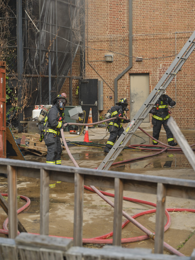 Feuerwehrleute in Helmen arbeiten daran, ein Gebäude Feuer zu löschen, während sie Schläuche halten, mit einem Metallzaun, Rohren, einem Container, einem Verkehrskegel und einem Baum im Hintergrund.
