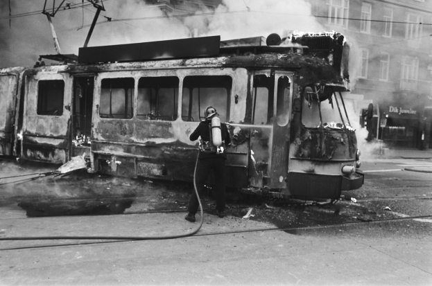 Schwarz-weiß-Foto eines Mannes in einem Helm, der eine Pfeife h├Ąlt und neben einer ausgebrannten Stra├čenbahn mit Rauch steht, vor einem Geb├Ąude mit Fenstern und Dr├Ąhten.