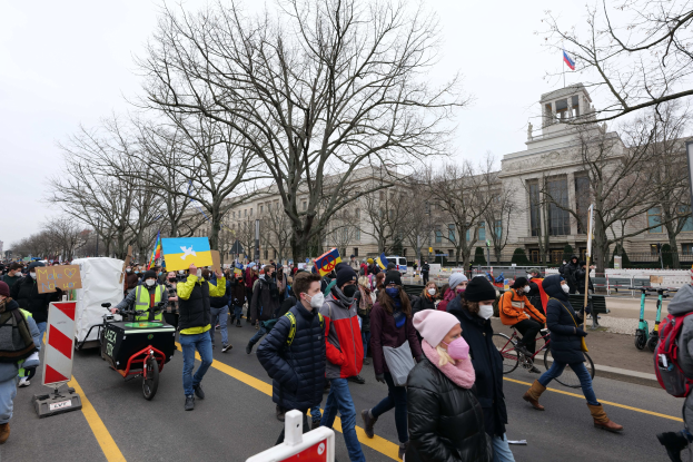Ein großer Protestmarsch mit Menschen, die eine Straße in Washington, D.C. entlanggehen, einige halten Schilder und andere fahren Fahrräder, mit Bäumen und einem klaren blauen Himmel im Hintergrund.