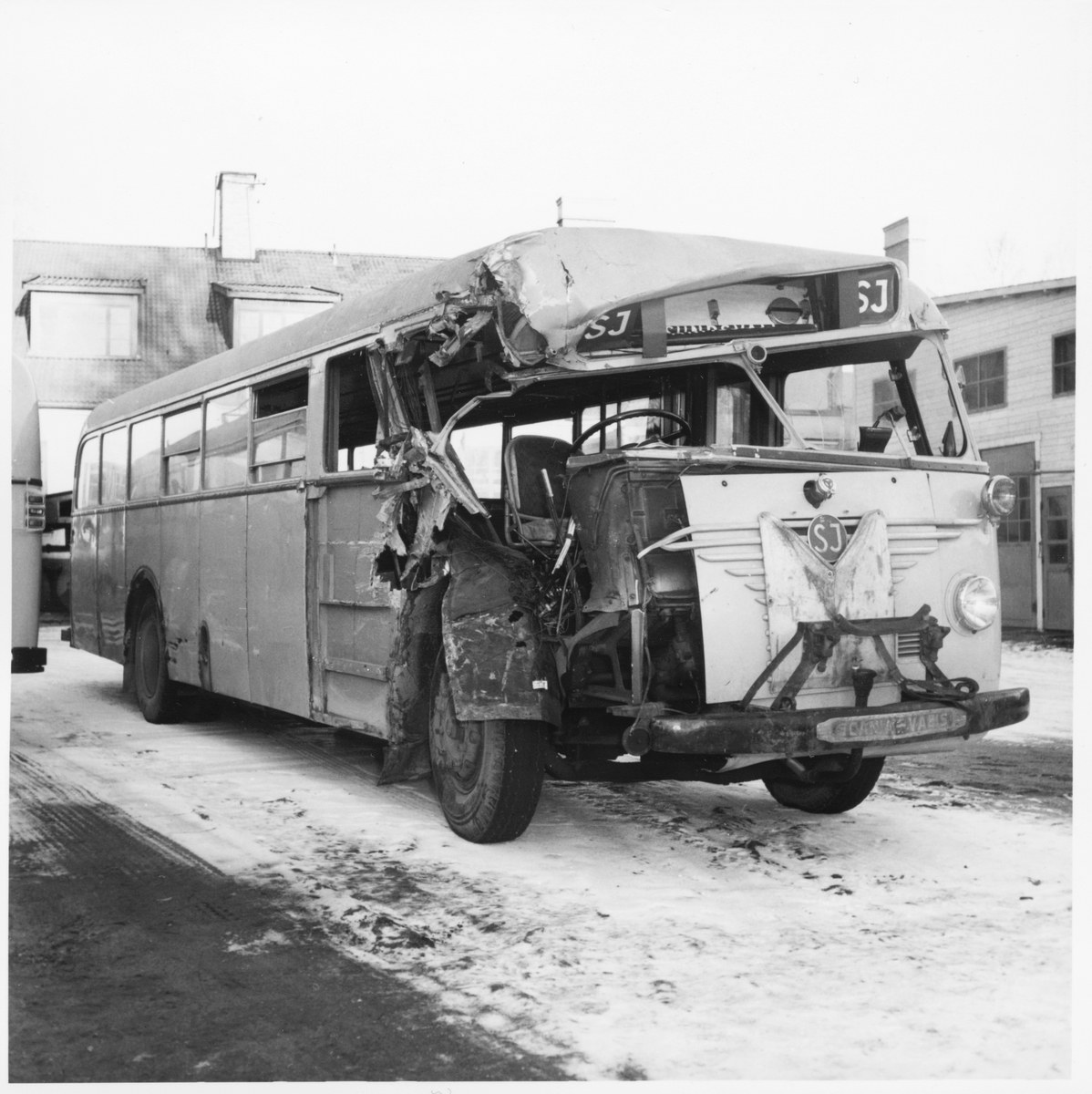Schwarz-weiß-Foto eines schwer beschädigten Busses am Straßenrand mit eingedellter Front und zersplitterten Fenstern vor einem Gebäudehintergrund.