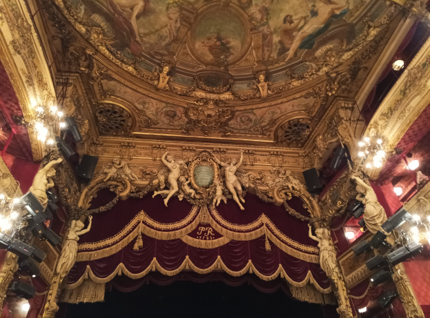 Theaterinnenraum mit roter Gardine, Kronleuchtern, Deckenplastiken, Lichtern und Gemälde, mit einer Bühne und einigen Personen unten.
