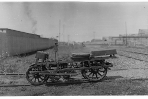 Ein altes Schwarz-Weiß-Foto eines Motorrads auf der Strecke, umgeben von Gras, Pflanzen, einer Wand, Pfosten, Gebäuden und einem Himmel im Hintergrund.