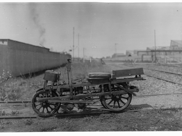 Ein altes Schwarz-Weiß-Foto eines Motorrads auf der Strecke, umgeben von Gras, Pflanzen, einer Wand, Pfosten, Gebäuden und einem Himmel im Hintergrund.