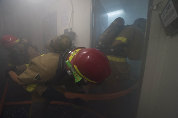 Feuerwehrleute in Schutzausrüstung mit Sauerstoffflaschen betreten einen raucherfüllten Raum, mit Rohren und einer Lampe im Hintergrund.