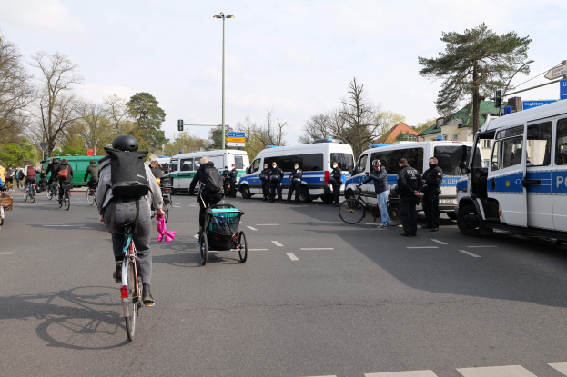 Eine Gruppe von Menschen auf Fahrrädern auf einer Straße mit Polizeiwagen, anderen Fahrzeugen, Bäumen, Straßenschildern, Gebäuden und einem bewölkten Himmel.