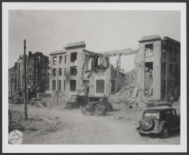 Schwarz-weißes Rahmenfoto einer Berliner Stadtstraße mit parkenden Autos vor einem schwer beschädigten Gebäude, wahrscheinlich von einer Bombenexplosion.