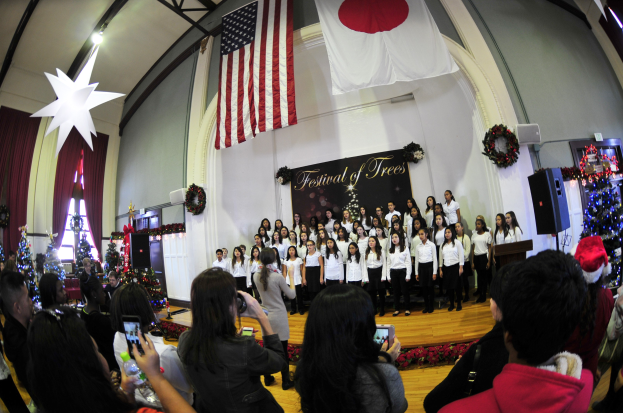 Eine Gruppe von Menschen auf einer Bühne vor einem Publikum, mit Weihnachtsdekoration, einem Rednerpult, Lautsprechern, einer Texttafel, Vorhängen, Flaggen und Deckenleuchten.