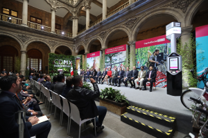 Eine Gruppe von Menschen sitzt in Stühlen und schaut auf einen Mann auf einer Bühne, der ein Mikrofon hält, mit Fahrrädern auf der rechten Seite und verschiedenen Hintergrundelementen wie Plakaten, Pflanzen, Säulen, Geländern, Lampen, Fenstern und einer Wand.