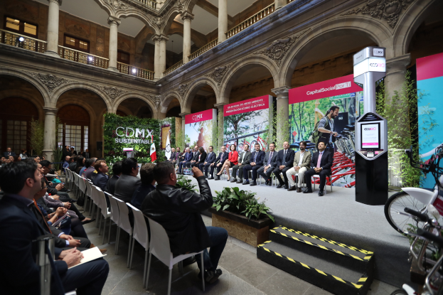 Eine Gruppe von Menschen sitzt in Stühlen und schaut auf einen Mann auf einer Bühne, der ein Mikrofon hält, mit Fahrrädern auf der rechten Seite und verschiedenen Hintergrundelementen wie Plakaten, Pflanzen, Säulen, Geländern, Lampen, Fenstern und einer Wand.