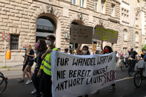 Eine Gruppe von Menschen nimmt an einer Protestaktion gegen die Anti-Kriegs-Bewegung in Berlin teil und marschiert mit Schildern, Bannern und Fahrrädern durch die Straßen, vorbei an einem Gebäude mit Fenstern, Bögen, Säulen und Skulpturen sowie Bäumen im Hintergrund.