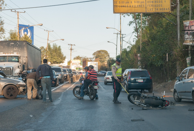 Gruppe von Menschen um ein verunglücktes Motorrad auf dem Seitenstreifen mit mehreren Fahrzeugen, darunter ein Lastwagen, im Hintergrund und Bäumen, Masten, Lichter, Schildern und Himmel.