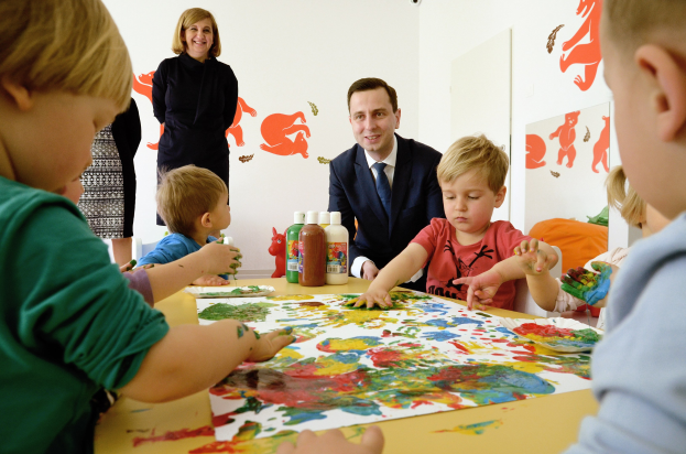 Kinder sind bei einer Malaktivität an einem Tisch mit Farbe an den Händen, Papieren und Flaschen beschäftigt, während eine Erwachsene im Hintergrund lächelt und Gemälde an der Wand zu sehen sind.