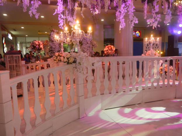 Hochzeitsempfang mit weißem Geländer, Blumenschmuck, Kronleuchtern an der Decke, Menschenansammlung, Säulen und Lichter im Hintergrund und ein Bildschirm auf der rechten Seite.
