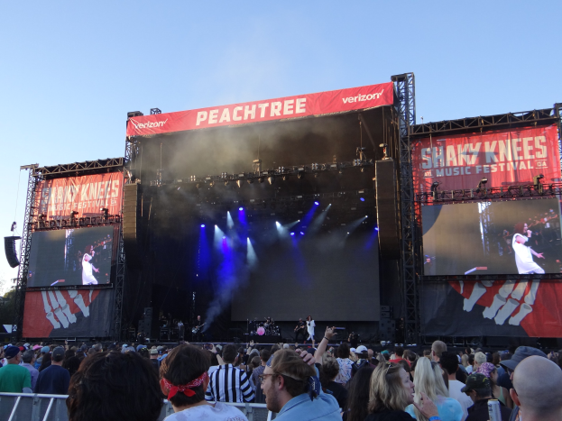 Eine große Menschenmenge steht vor einer hell erleuchteten Bühne beim Shaky Knees Music Festival, auf der Musiker unter Bannern spielen, mit einem Geländer im Vordergrund und einem klaren blauen Himmel im Hintergrund.