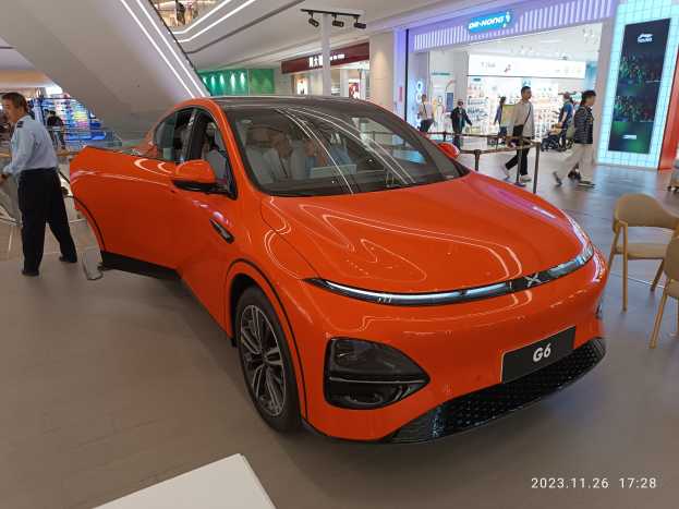 Oranger Elektroauto in einer Ausstellungshalle mit einer Gruppe von Menschen drumherum, umgeben von Stühlen, einem Wagen, Geschäften, Namensschildern und Lichtern im Hintergrund.