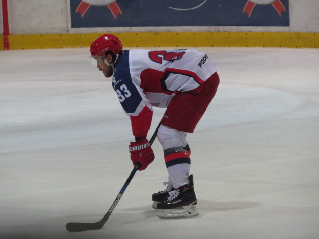Eishockey-Spieler in voller Ausrüstung auf dem Eis mit einem Stock, der einer Wand mit Banner im Hintergrund zugewandt ist.