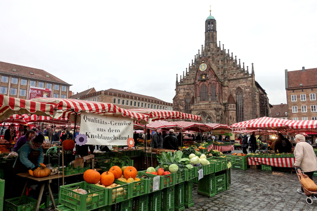 Ein belebter Markt in Nürnberg, Deutschland, mit Ständen, auf denen verschiedene Obst- und Gemüsesorten angeboten werden, Menschen mit Taschen und aufgestellte Zelte im Hintergrund, mit Gebäuden und einem Uhrenturm.