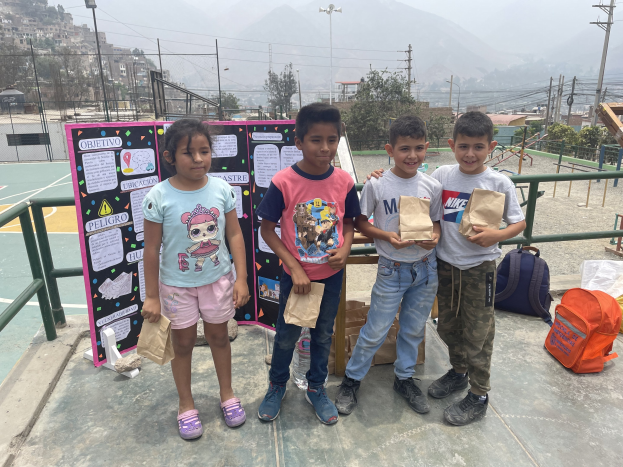 Gruppe von Kindern auf einem Basketballfeld, die Papierbeutel halten und Gegenstände auf dem Boden rechts und eine Tafel mit Text links haben, vor einem Hintergrund aus Zaun, Pfosten, Straßenlaternen, Bäumen, Gebäuden, Hügeln und Himmel.