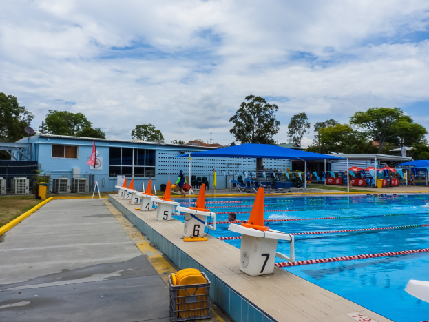 Ein großes Freibad mit schwimmenden Menschen, umgeben von Bahnmarkierungen, Verkehrskegeln, Stühlen, Sonnenschirmen, einem Gebäude mit Fenstern, einer Flagge, Bäumen und einem bewölkten Himmel.