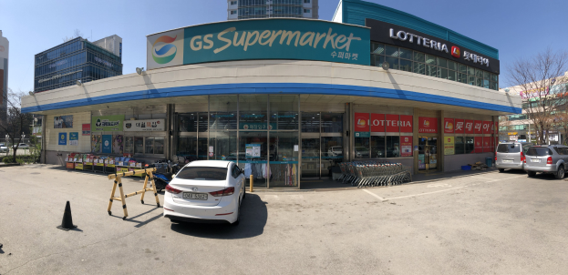 Ein Auto vor einem "GS Supermarkt" mit anderen Fahrzeugen, Verkehrskegeln und verstreuten Gegenständen in der Nähe, vor dem Hintergrund von Gebäuden, Bäumen und einem klaren blauen Himmel.
