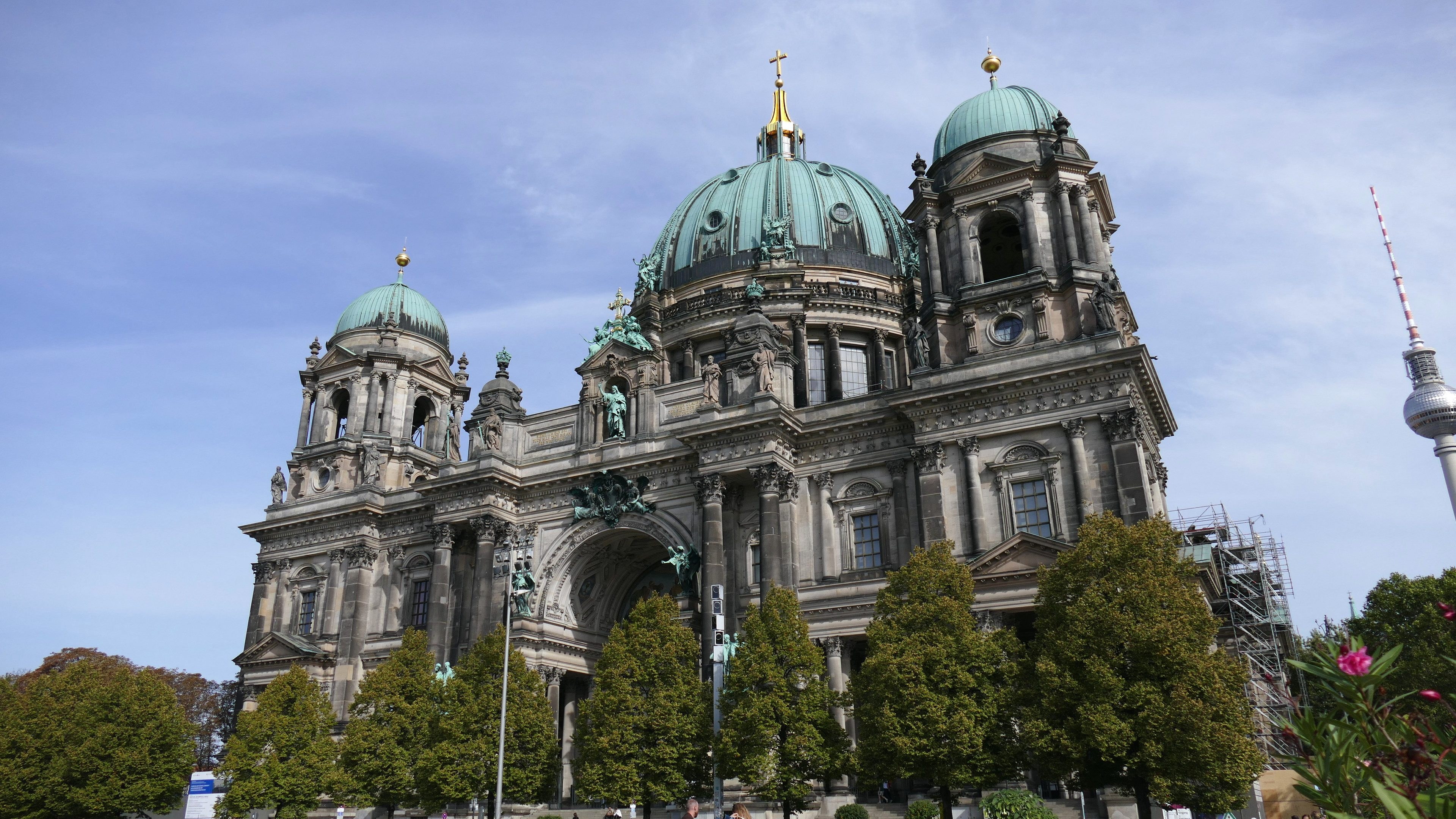 Der Berliner Dom, eine große Kathedrale mit detaillierter Architektur, steht prominent in Berlin, Deutschland, mit Menschen, Bäumen und einem Turm im Vordergrund unter einem bewölkten Himmel.