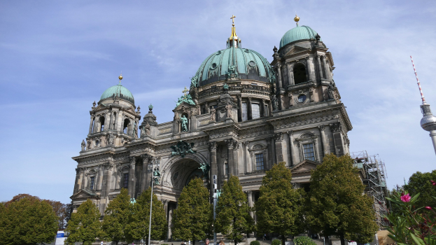 Der Berliner Dom, eine große Kathedrale mit detaillierter Architektur, steht prominent in Berlin, Deutschland, mit Menschen, Bäumen und einem Turm im Vordergrund unter einem bewölkten Himmel.