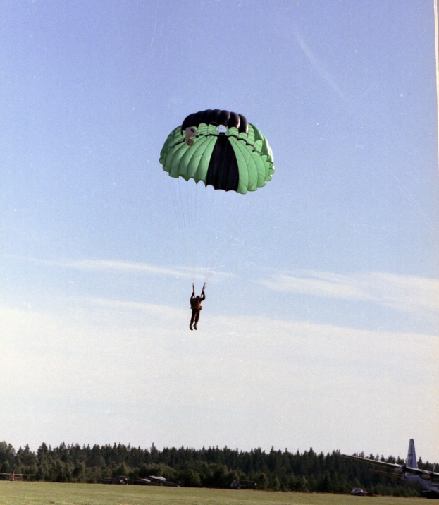 Eine Person beim Paragleiten in der Luft mit einem Fallschirm, mit einem Flugzeug rechts im Bild, vor dem Hintergrund von Gras, Bäumen und bewölktem Himmel.