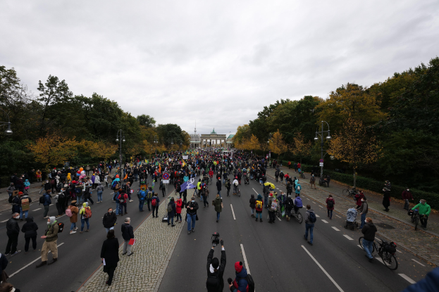 Eine große Gruppe von Menschen, die eine von Bäumen gesäumte Straße in Berlin entlanggehen, einige halten Kameras, während sie an einer Protestdemo unter einem klaren Himmel teilnehmen.