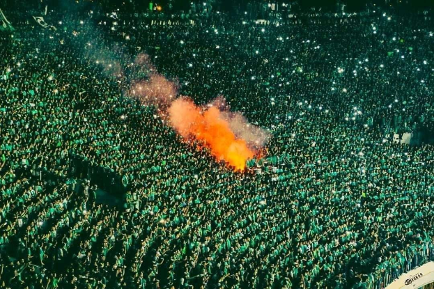 Luftaufnahme einer großen Menge, die in einem Stadion steht, mit einem zentralen Feuer, das die Umgebung erhellt.