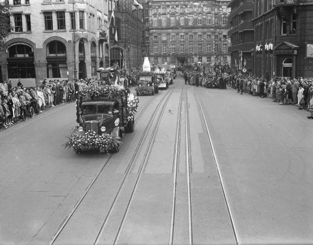 Ein Schwarz-Weiß-Foto eines Umzugs auf einer Stadtstraße mit Fahrzeugen, Zuschauern auf beiden Seiten und Gebäuden mit Fenstern, Laternenmasten und einem Baum im Hintergrund.