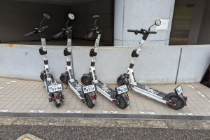 Drei schwarze und weiße E-Scooter mit Nummern und Text nebeneinander auf einem Gehweg vor einem Gebäude mit Glastür und Wand im Hintergrund geparkt.