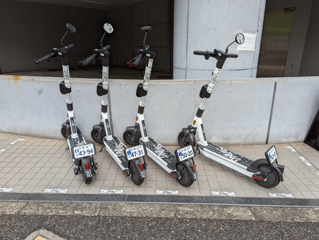 Drei schwarze und weiße E-Scooter mit Nummern und Text nebeneinander auf einem Gehweg vor einem Gebäude mit Glastür und Wand im Hintergrund geparkt.