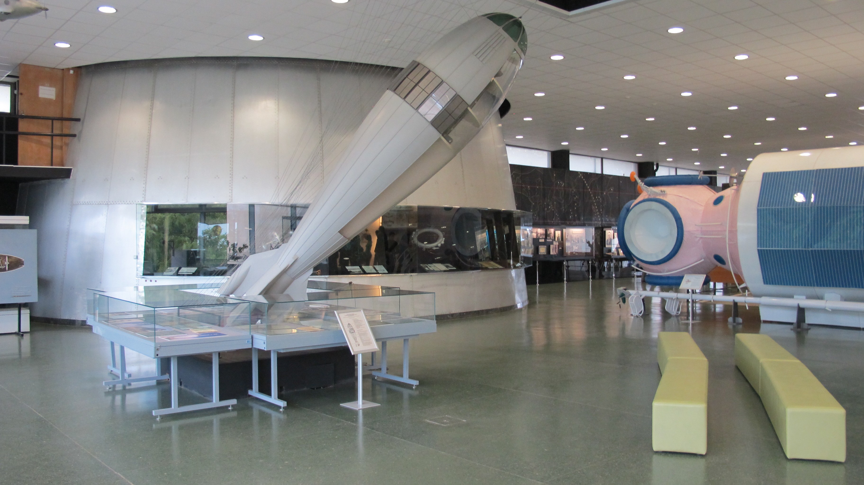 Großer Raum mit verschiedenen Weltraumgeräten, einschließlich eines Raketen auf der rechten Seite, Sitzbereiche, eine Tafel mit Text auf einem Tisch, Zaun, Wände, Deckenleuchten und ein Fenster.