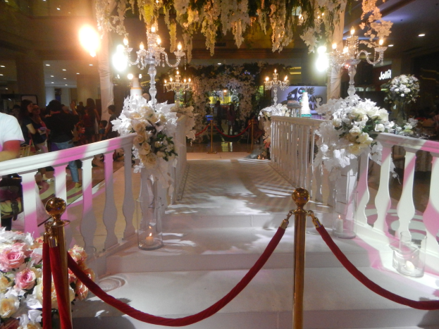 Eine Hochzeitszeremonie in einem Einkaufszentrum mit weißem Teppich, dekorativen Geländern, Blumenarrangements, Kronleuchtern und Deckenbeleuchtung, bei der viele Menschen anwesend sind.