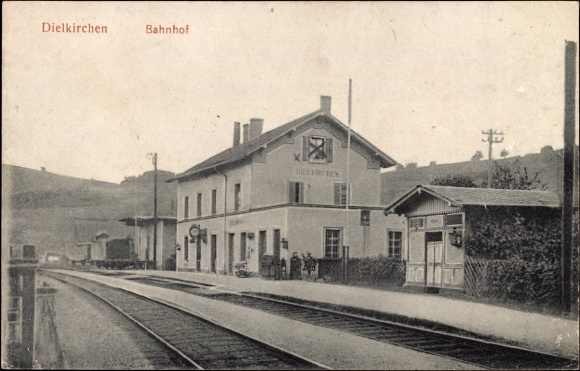 Schwarzes und weißes Foto einer Bahnstation in Dielkirchen, Deutschland, mit einem Zug auf den Gleisen, umgeben von Häusern, Strommasten, Bäumen und Menschen, vor Bergen und einem klaren Himmel.