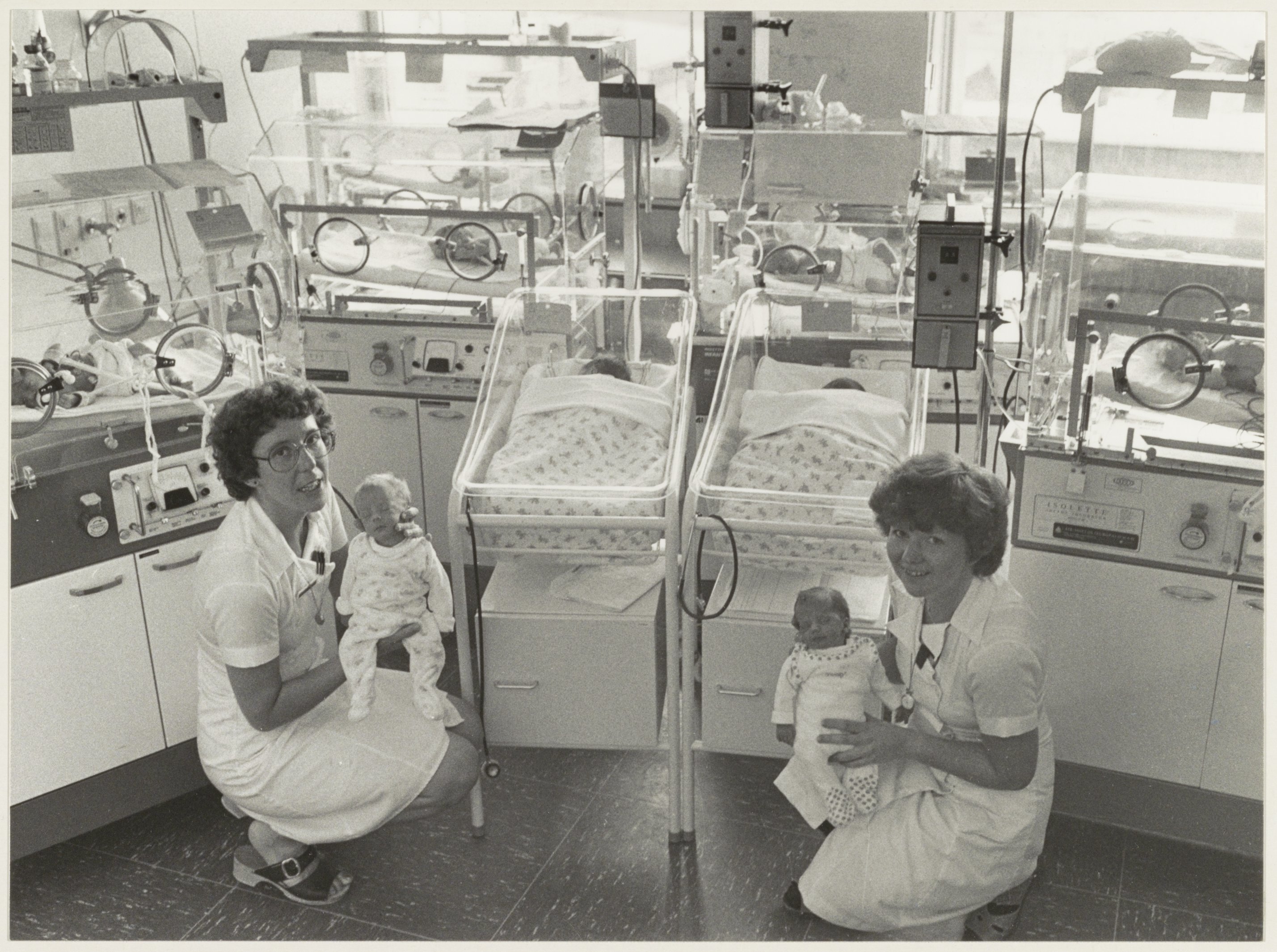 Ein Schwarz-Weiß-Foto von zwei Frauen in einem Krankenhauszimmer, jede hält ein Baby, mit Inkubatoren und medizinischer Ausrüstung im Hintergrund.