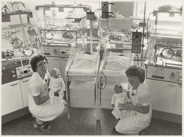 Ein Schwarz-Weiß-Foto von zwei Frauen in einem Krankenhauszimmer, jede hält ein Baby, mit Inkubatoren und medizinischer Ausrüstung im Hintergrund.