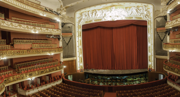 Großer Auditoriumsinnenraum mit roter Gardine, Sitzreihen, Geländern, Beleuchtung, Wandskulpturen und Bühnenobjekten, ähnlich einem Konzertsaal.
