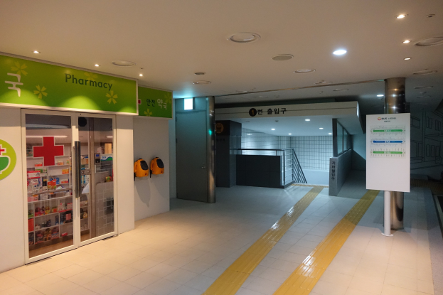 Eingang einer Apotheke mit einem "Apotheke"-Schild links, einer Texttafel rechts und beleuchteten Lampen über dem Dach.