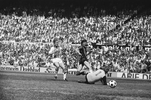 Schwarzes und weißes Foto von Männern, die auf einem Feld Fußball spielen, mit Zuschauern im Hintergrund und einer "Bundesliga 1978-79 Ajax 1-1 Hannover 96"-Fahne.