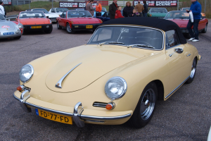 Ein gelber Porsche 356 Speedster auf einem Parkplatz mit Menschen in der Nähe, vor einer Umzäunung, Bannern, Bäumen, Gebäuden und einem klaren Himmel.