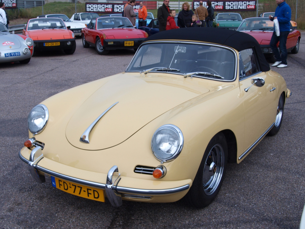Ein gelber Porsche 356 Speedster auf einem Parkplatz mit Menschen in der Nähe, vor einer Umzäunung, Bannern, Bäumen, Gebäuden und einem klaren Himmel.
