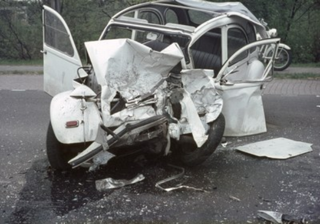Schwerbeschädigte Autos am Straßenrand mit Frontalaufprall, Bäume und ein Gebäude im Hintergrund, weiteres Fahrzeug in der Ferne sichtbar.
