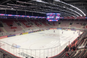 Eishockeyspiel in einer großen Arena mit Zuschauern, ein Eisring umgeben von einem Zaun, ein Display, das "St. Louis Blues vs Detroit Red Wings" anzeigt, Metallrahmen, Deckenleuchten und ein Dach.
