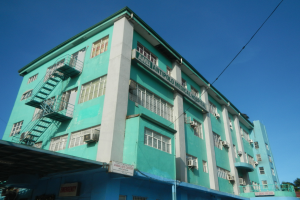 Ein großes grünes Gebäude mit vielen Fenstern, Klimaanlagen, Treppen, Geländern, Namensschildern, Strommasten, Stromkabeln, Fahrzeugen auf der Straße, Bäumen und einem klaren blauen Himmel.